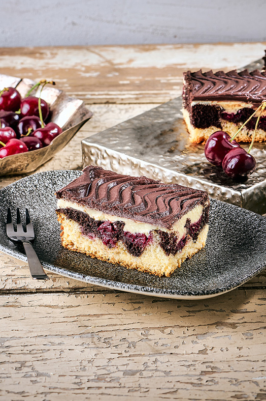 Ein Stück Donauwelle Kuchen mit Schokoladenglasur und Kirschen auf einem schwarzen Teller.
