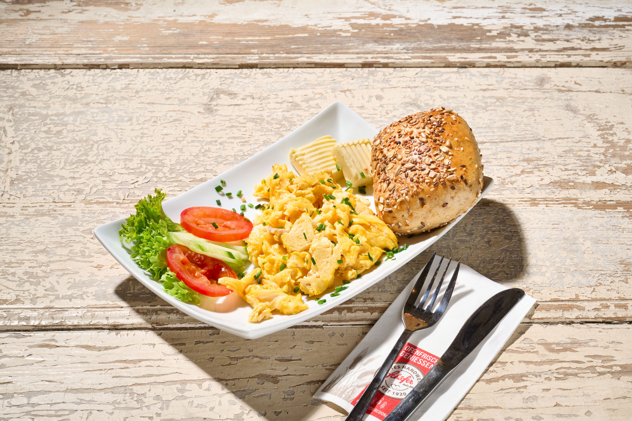 Herzhaftes Rührei-Frühstück Rustikaler Teller mit Rührei, Butter, Salat und einem Körnerbrötchen auf hellem Holz