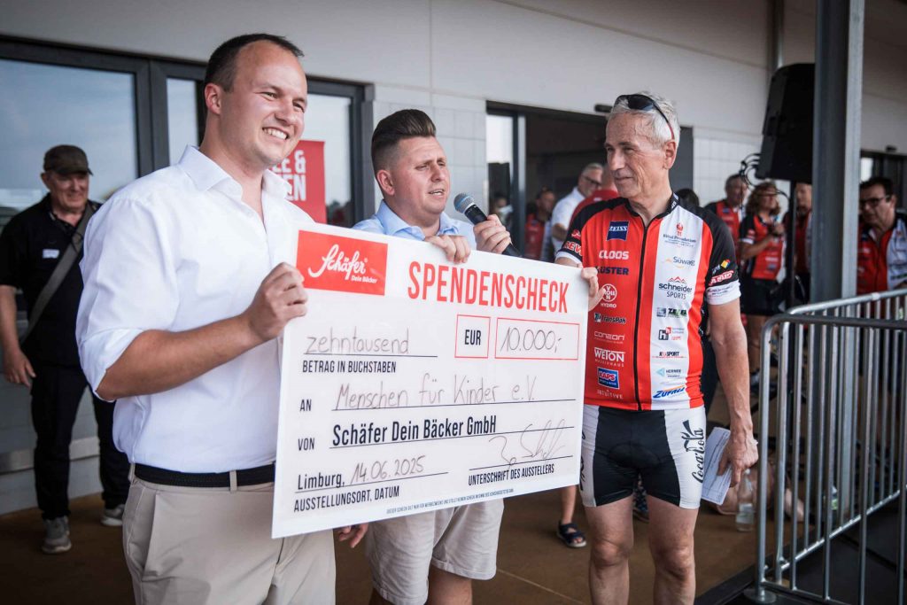 Drei lächelnde Personen stehen auf einer Bühne, zwei von ihnen halten gemeinsam einen überdimensionalen Spendenscheck über 10.000 € mit dem Logo „Schäfer Dein Bäcker“. Einer der Männer spricht in ein Mikrofon, im Hintergrund sind Zuschauer und Radfahrer zu sehen.