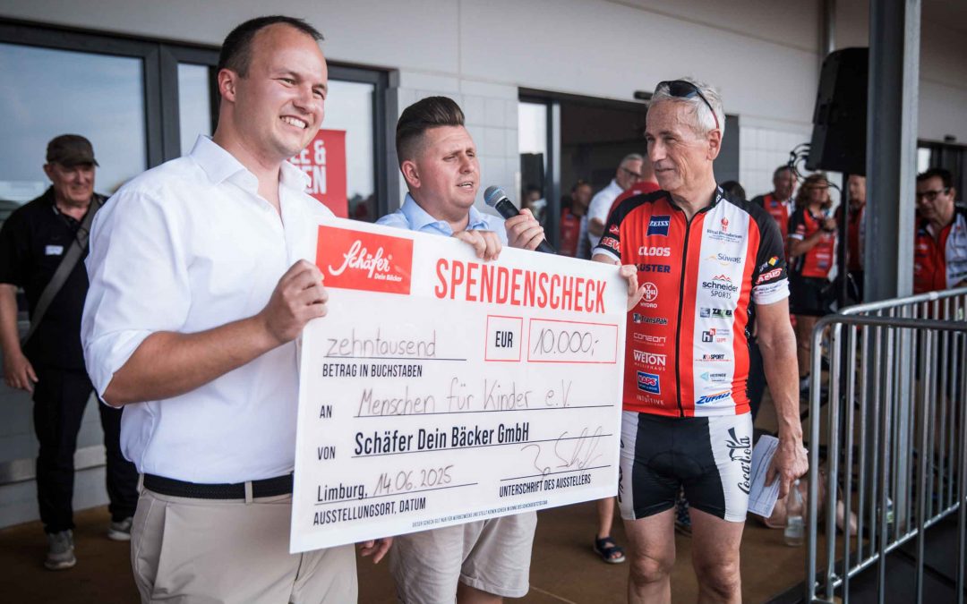 Drei lächelnde Personen stehen auf einer Bühne, zwei von ihnen halten gemeinsam einen überdimensionalen Spendenscheck über 10.000 € mit dem Logo „Schäfer Dein Bäcker“. Einer der Männer spricht in ein Mikrofon, im Hintergrund sind Zuschauer und Radfahrer zu sehen.