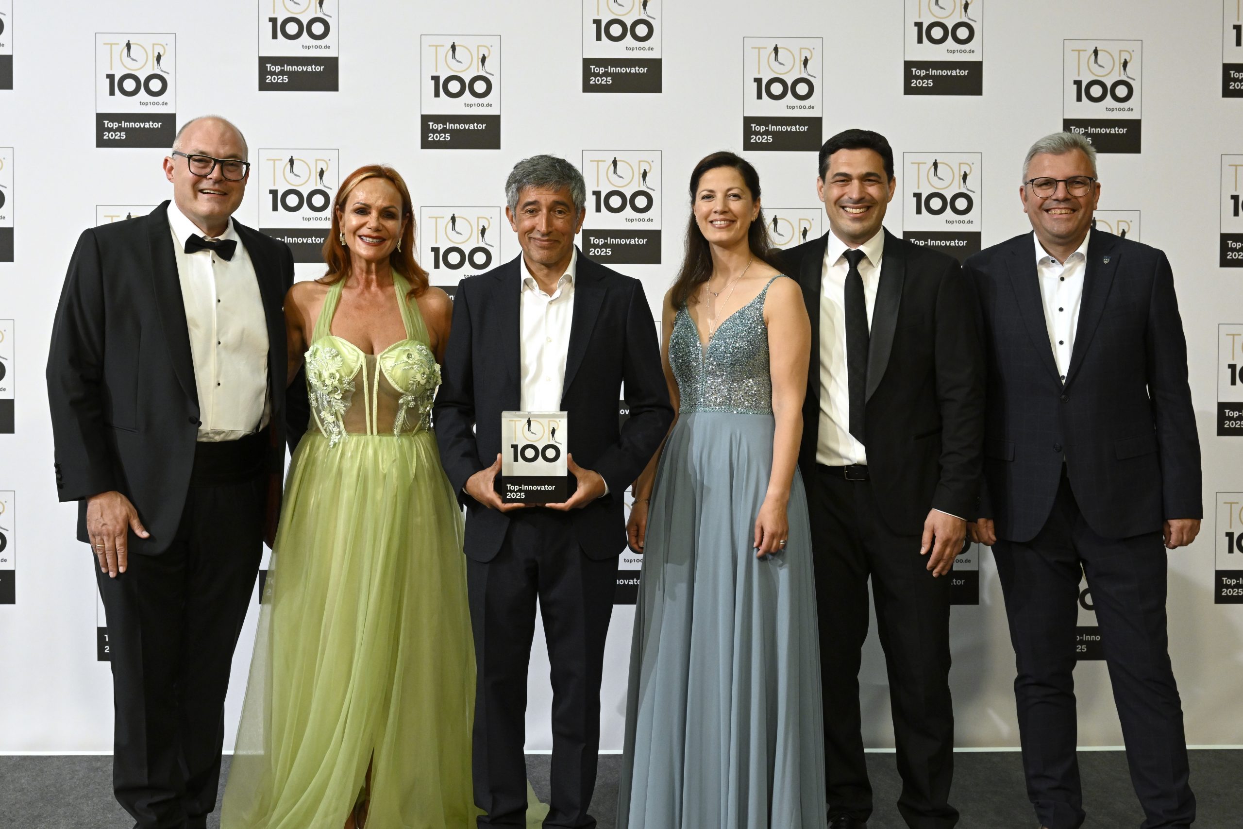Sechs Personen in festlicher Abendgarderobe vor einer Fotowand mit „TOP 100 Top-Innovator 2025“-Logos, der in der Bildmitte stehende Mann hält das „TOP 100“-Trophäenschild.