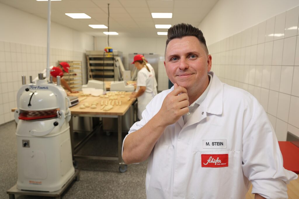 Ein Herr ist in der Backstube von Schäfer Dein Bäcker, in Arbeitskleidung mit Namensschild mit der Beschriftung M. Stein, während im Hintergrund Mitarbeitende Teig vorbereiten.