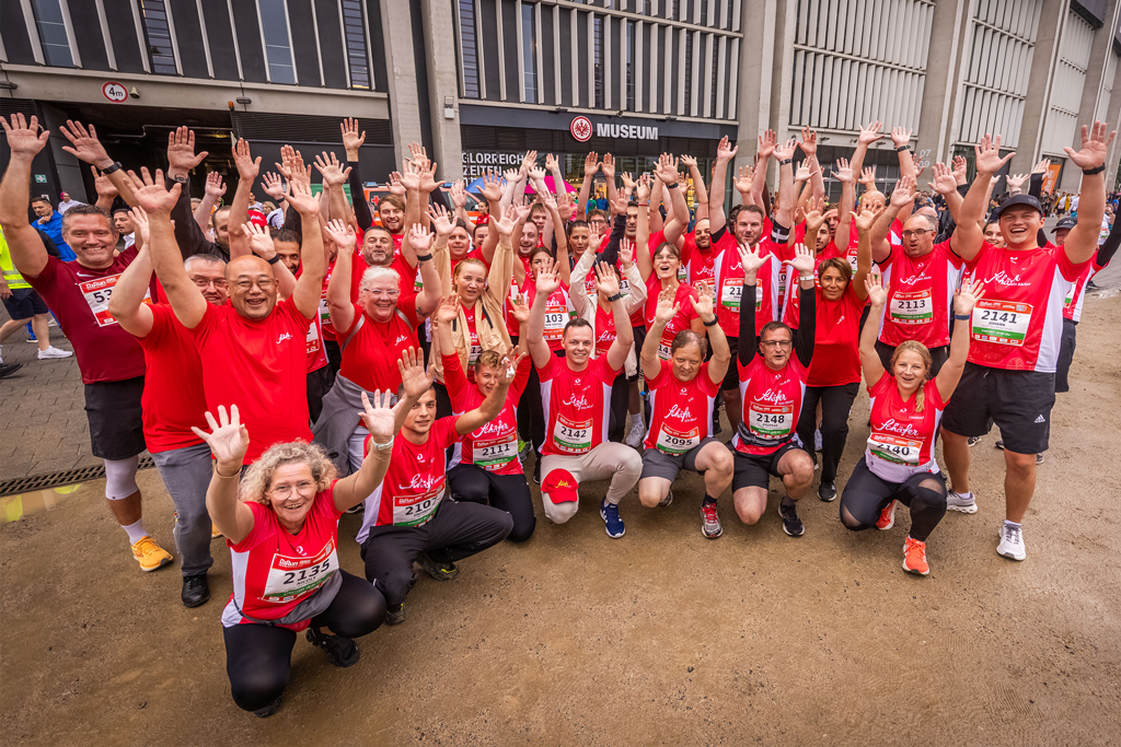 Großes Teamfoto von Läuferinnen und Läufern in roten „Schäfer Dein Bäcker“-Shirts, die lachend die Hände in die Luft heben — Gruppenbild beim B2Run Frankfurt 2025.