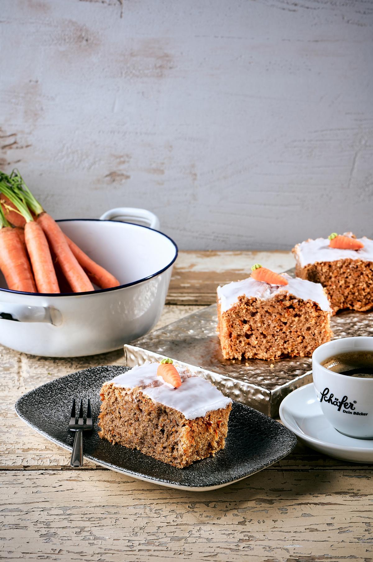 K1600_70 Rueblikuchen 012 Stück saftiger Karottenkuchen mit Zuckerglasur und Marzipankarotte neben Kaffee