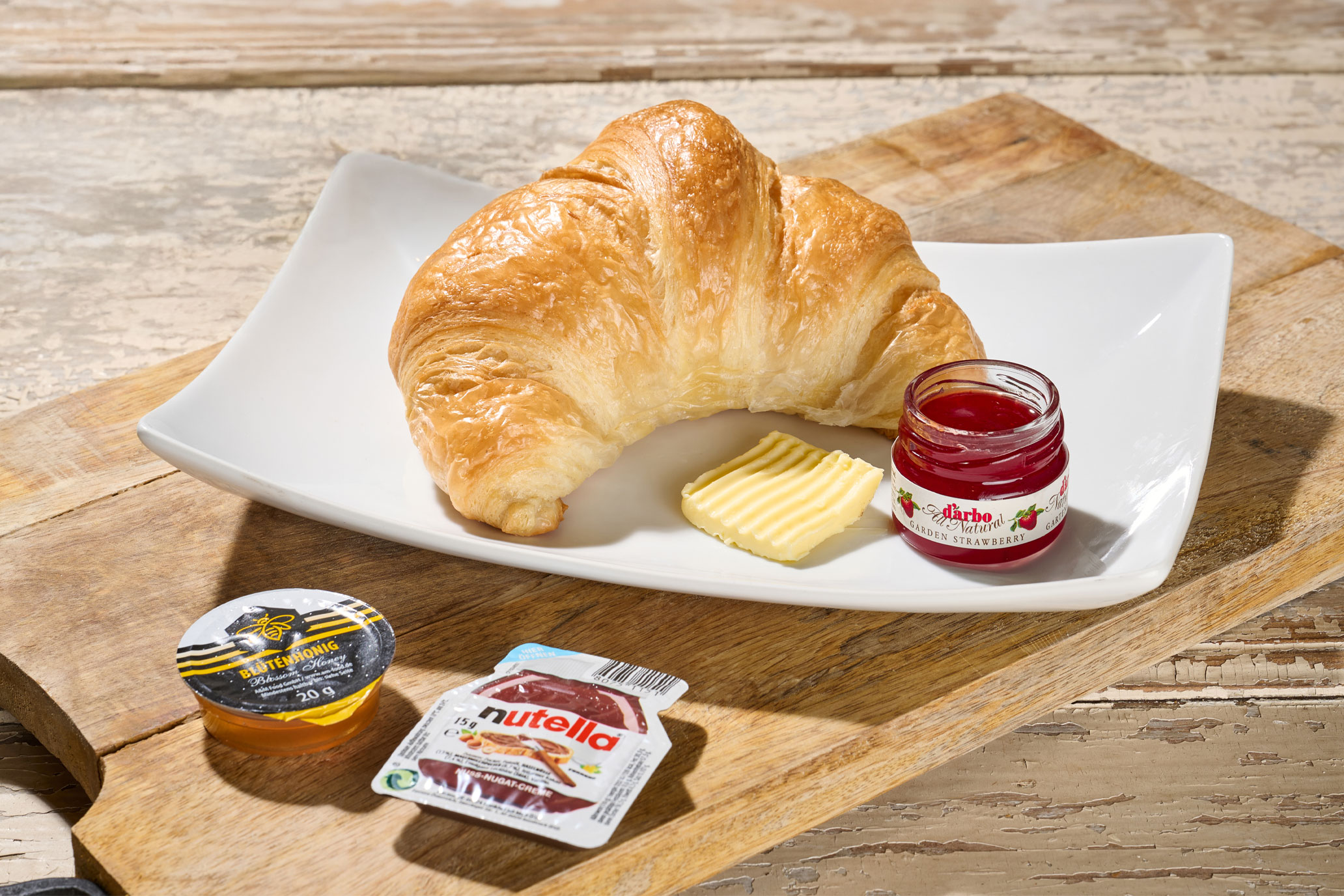 Buttriges Croissant mit Butter, Erdbeermarmelade, Honig, Nutella und einer Tasse Cappuccino auf Holzbrett