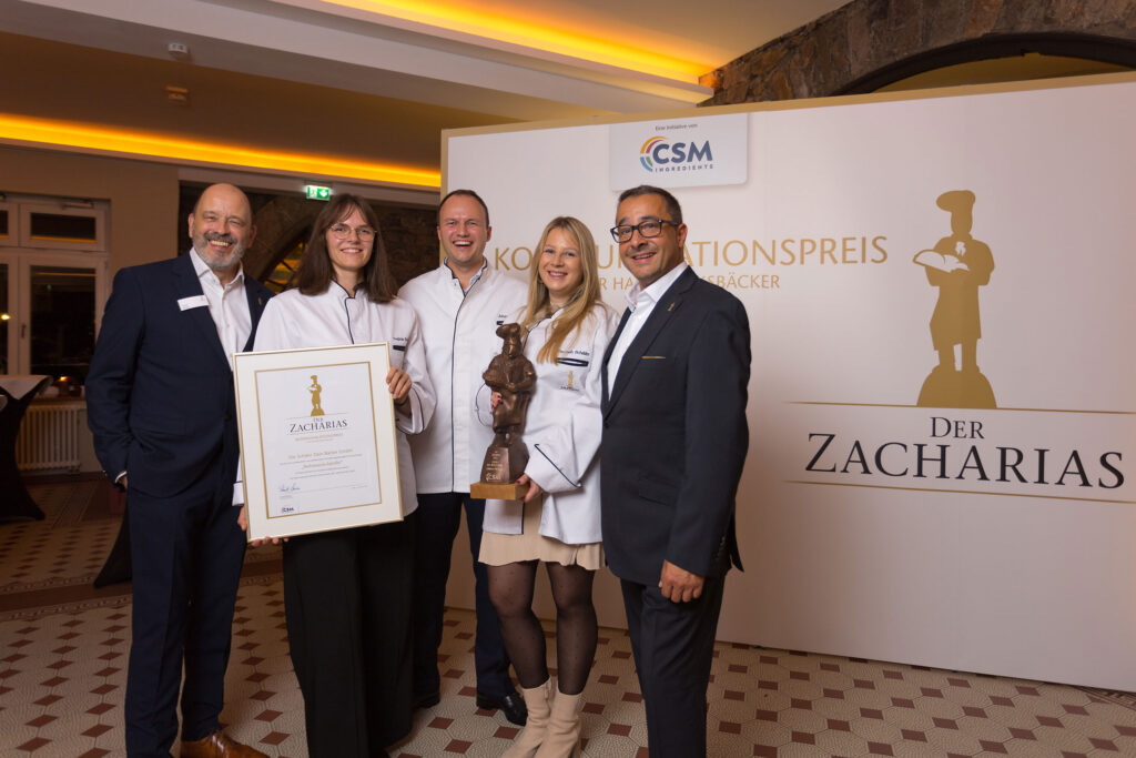 Fünf Personen halten eine Urkunde und eine Zacharias-Trophäe vor einer Fotowand.